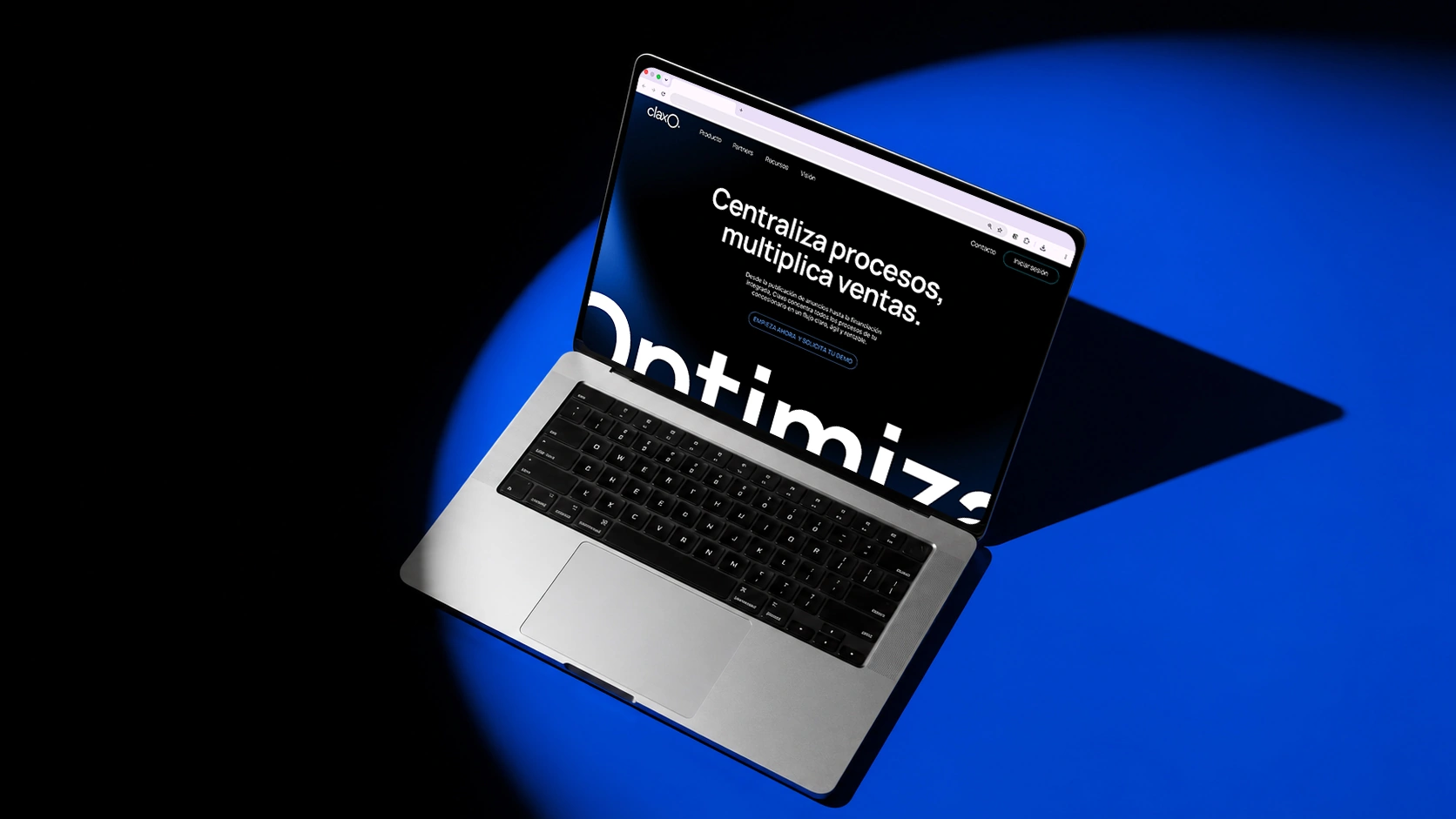 Un ordenador portátil, colocado sobre una superficie azul, muestra en la pantalla una página web con las palabras «Centraliza procesos, multiplica ventas», parcialmente iluminada por un foco.