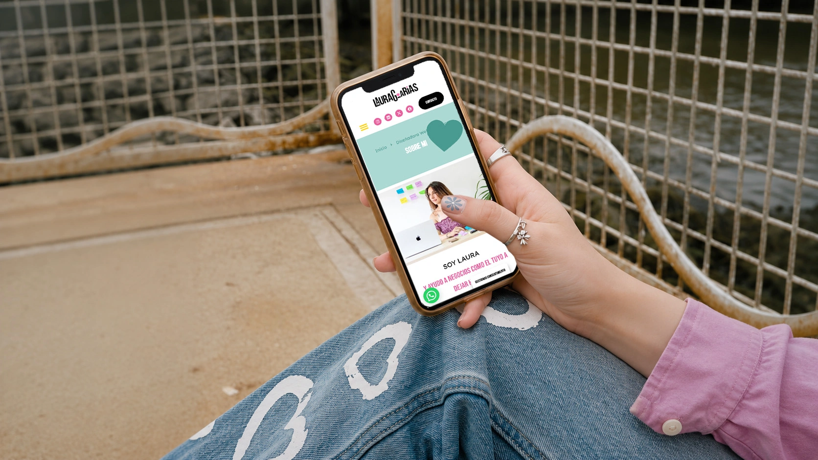 Persona sentada al aire libre con un smartphone en la mano en el que se ve una aplicación de citas; lleva vaqueros azules con motivos blancos y una camiseta rosa.