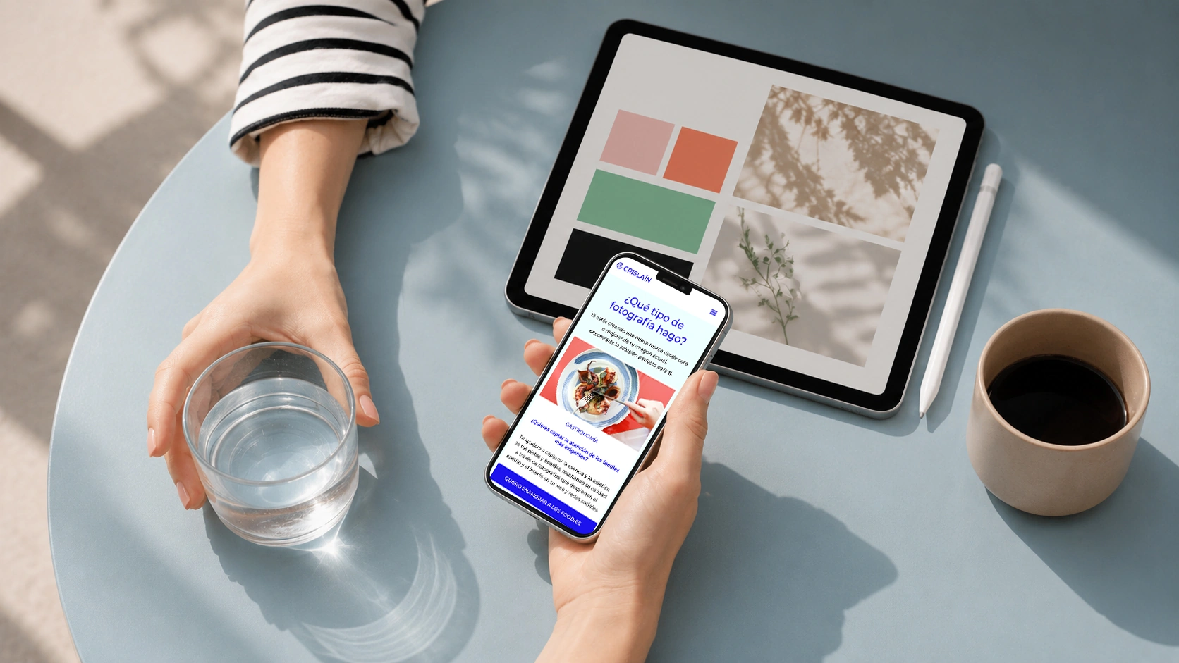 Persona que sostiene un vaso de agua y un smartphone, junto a una tableta en la que se ve una paleta de colores y una taza de café sobre una mesa redonda azul.